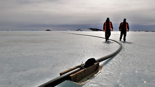 Two expeditioners walking along the fuel line with ship in background
