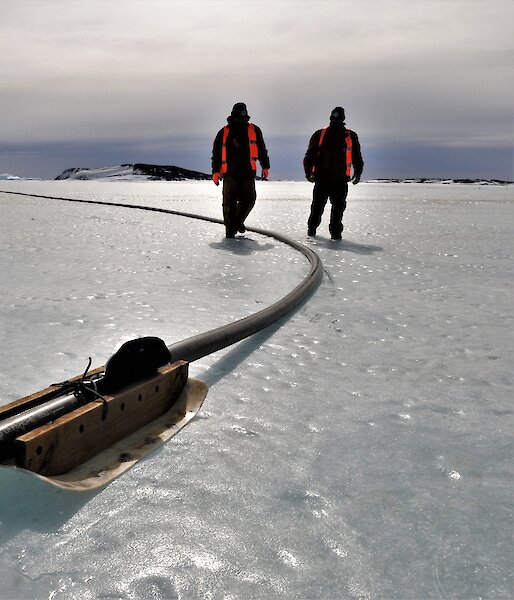 Two expeditioners walking along the fuel line with ship in background