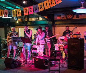 Band group in action bathed in pink light amongst speakers and music stands