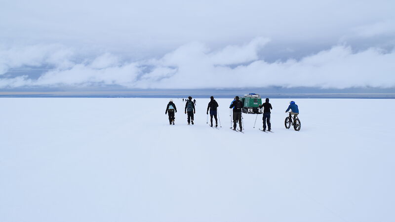 Snow covered ground to horizon (mid frame), grey sky with white band of cloud just above. Group of six expeditioners, mid frame, five skiing one on a bike, with green Hagglands just in front