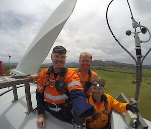 Selfie of three electricians on top of wind turbine with green verdant flat land stretching out below to distant mountain ranges