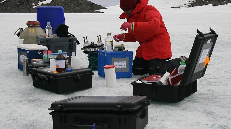Expeditioner kneeling on ice in front of boxes of scientific equipment