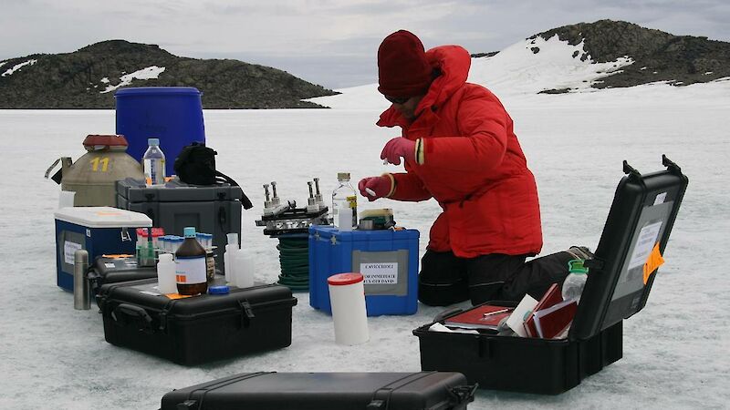 Expeditioner kneeling on ice in front of boxes of scientific equipment