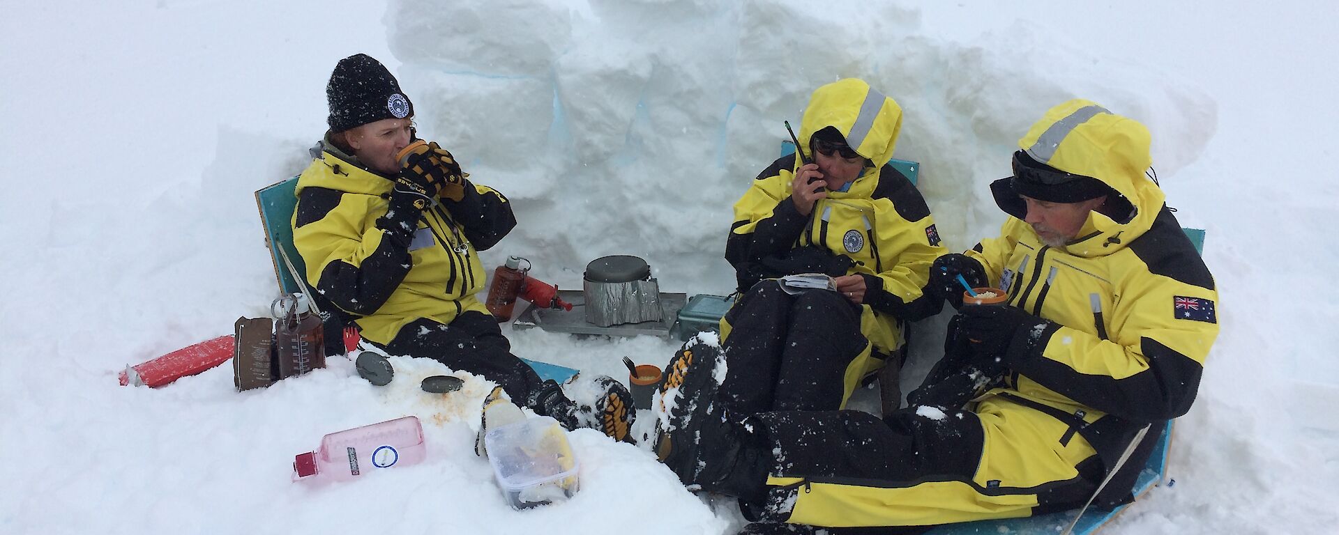 Three expeditioners sitting in a dug out snow hollow eating from mugs with middle expeditioner on the radio reading from a notebook