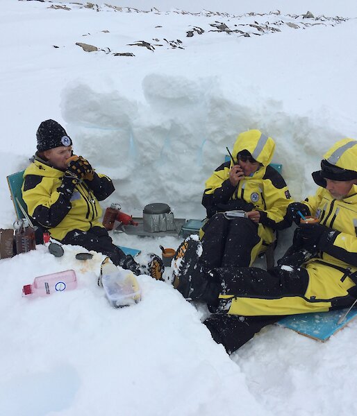 Three expeditioners sitting in a dug out snow hollow eating from mugs with middle expeditioner on the radio reading from a notebook