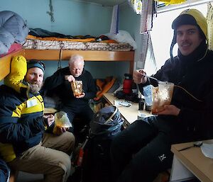 Three men inside a small field hut eating from small plastic bags