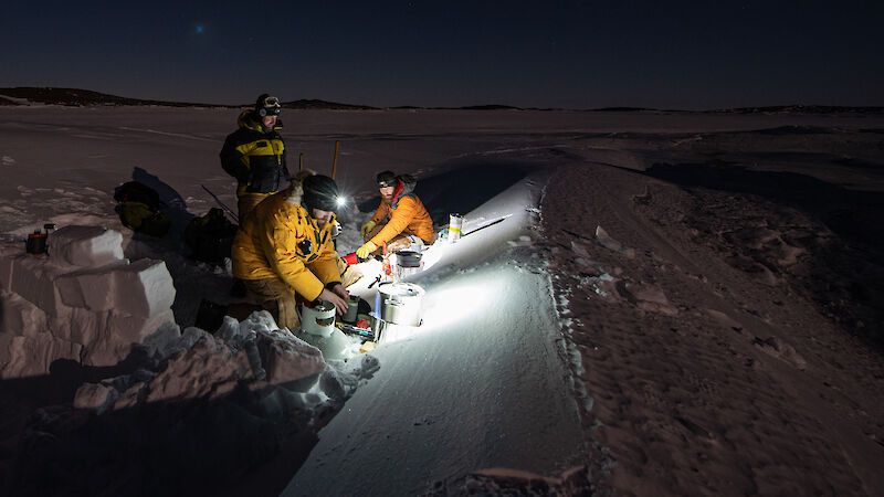 A twilight photo of 3 expeditioners sit in the snow with cooking pots and camp stoves.