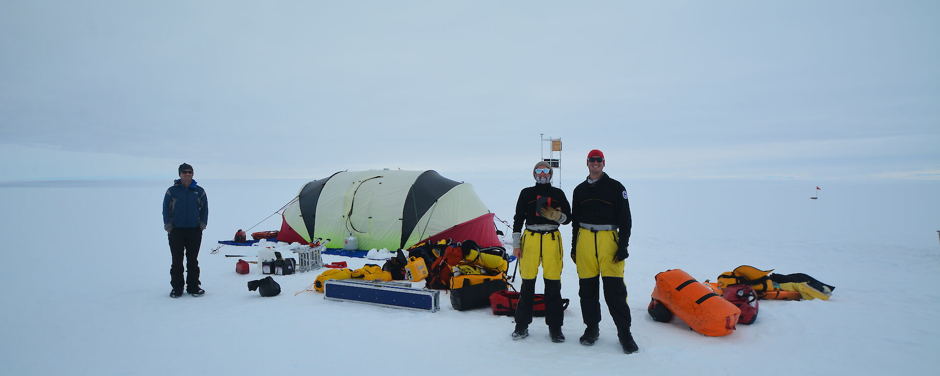 A tent, expeditioners and field equipment littering the snow on flat expanse of glacier