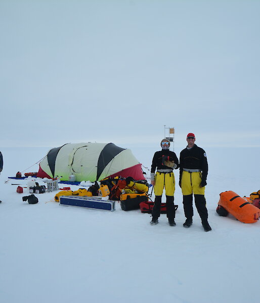 A tent, expeditioners and field equipment littering the snow on flat expanse of glacier