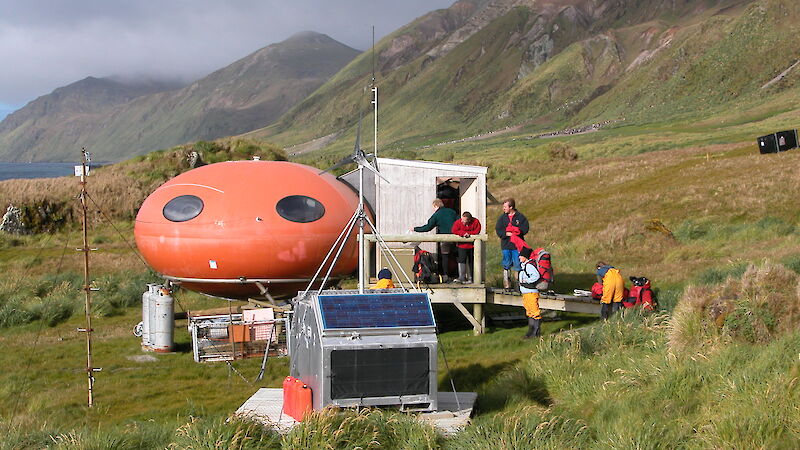 Red round hut set on the grassy slope of the island with expeditioners standing outside.