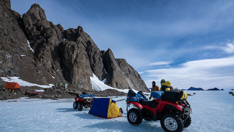 Small field hut nestled at base of steep rock mountain with quad bikes parked nearby.