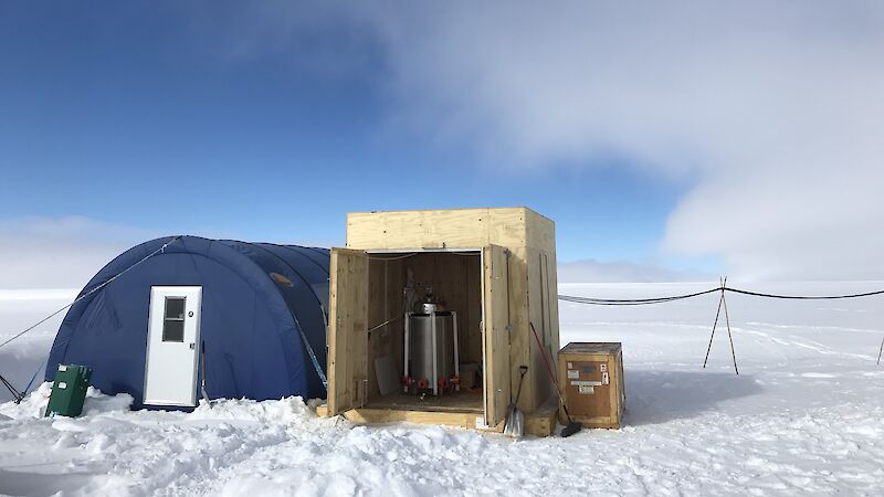 Blue semi-circular Weatherhaven tent and metal generator in wooden container stand on snowy ground.