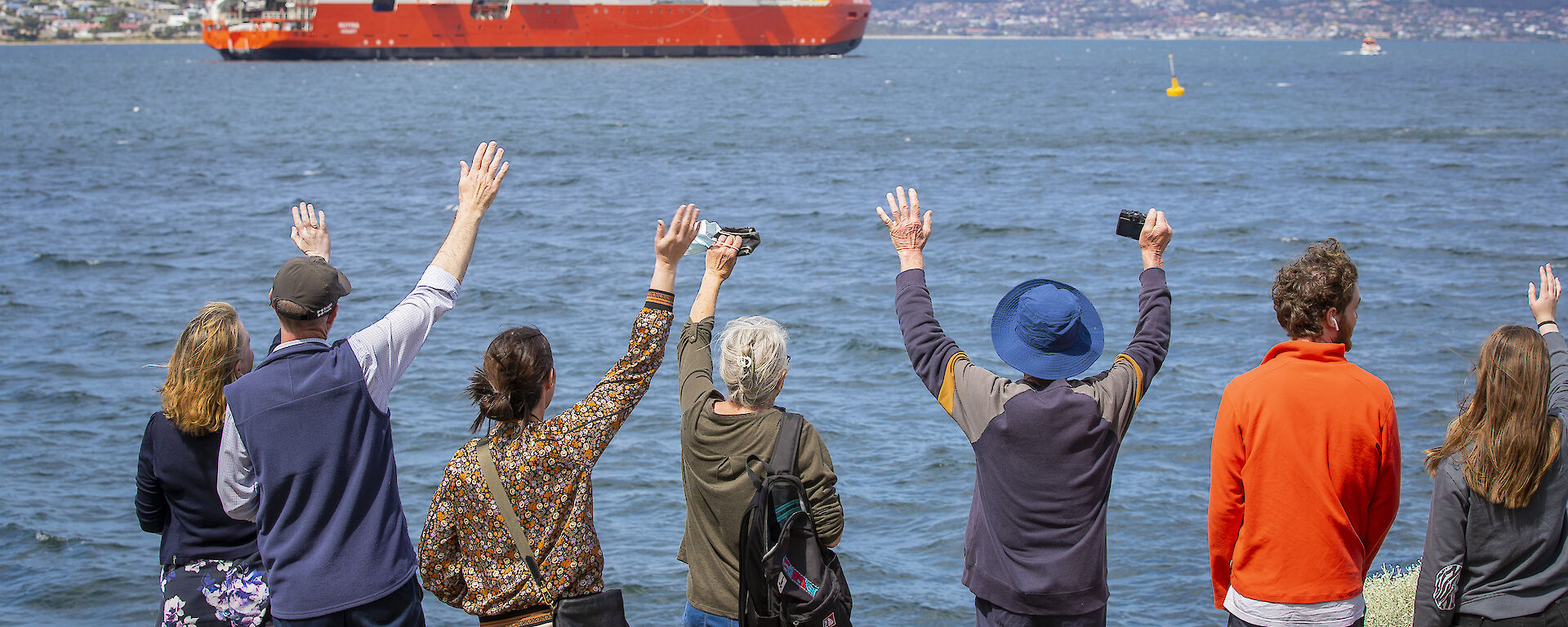 An orange and white ship sales away from the wharf. Onlookers wave it goodbye.