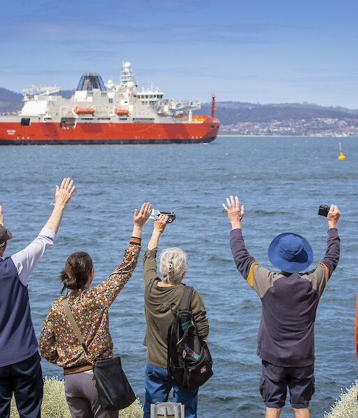 An orange and white ship sales away from the wharf. Onlookers wave it goodbye.