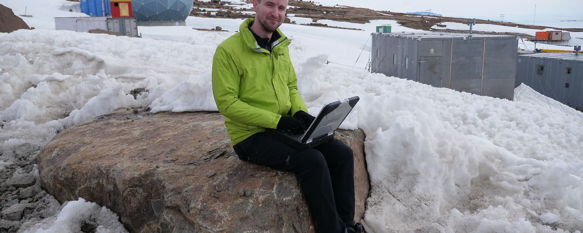 An expeditioner sitting on a rock at Mawson with a laptop. The ANARESAT radome and some of the heritage station buildings are in the background.