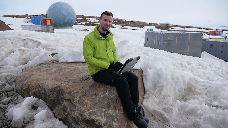 An expeditioner sitting on a rock at Mawson with a laptop. The ANARESAT radome and some of the heritage station buildings are in the background.