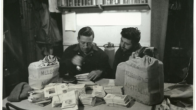 A black and white image of two expeditioners behind a desk covered with stacks of letters.