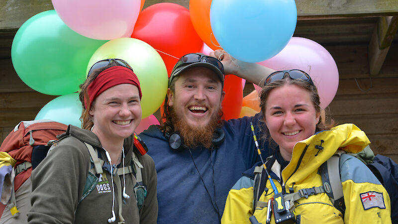 Expeditioners wearing their wet weather gear and carry-on back packs arrive back at station, and an expeditioner greets them with balloons to decorate the mess for a birthday party.