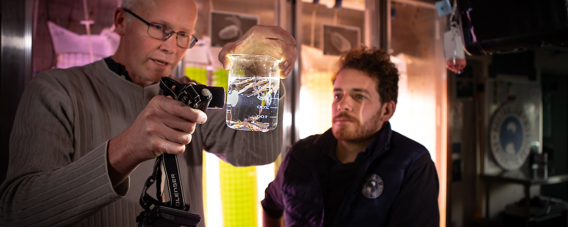 2 scientists stand in an aquarium lab. They hold a light up to a beaker of water in which small organisms are swimming.