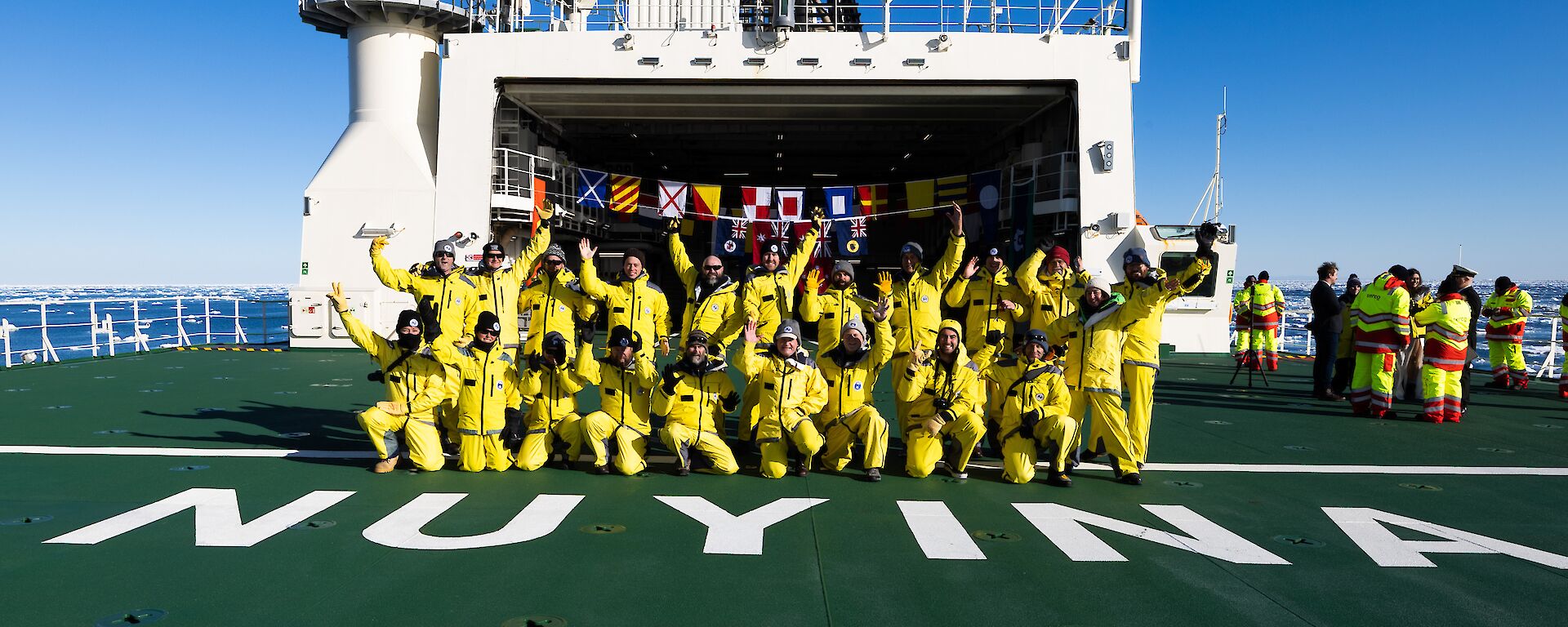 A large group of expeditioners dressed in yellow, pose for a group photo on the deck of a ship.