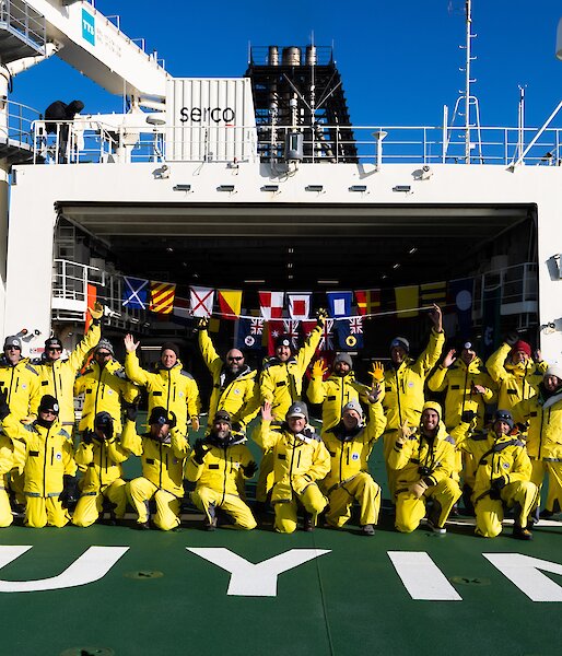 A large group of expeditioners dressed in yellow, pose for a group photo on the deck of a ship.