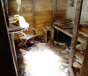 An old wooden chair sits in a small room by a hard bunk bed. Books on a shelf are frozen and drip ice.