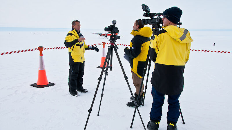 A man in cold weather clothing being interviewed on the ice by a journalist and camera operator.