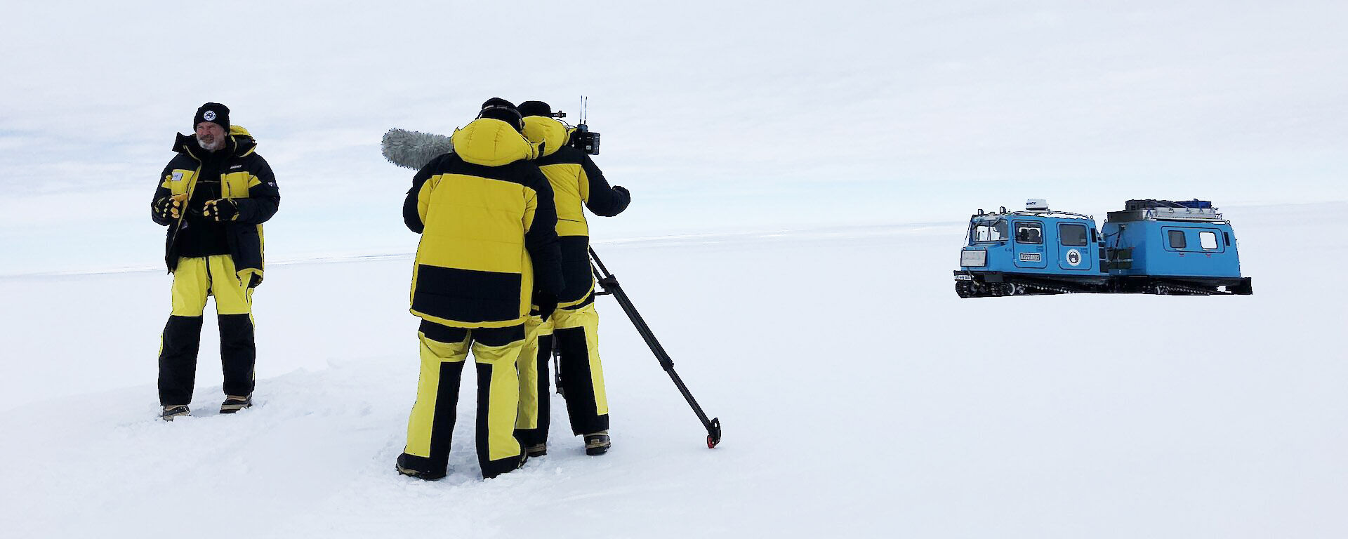 Yellow survival suited film crew on the ice with blue Hagglunds in near distance