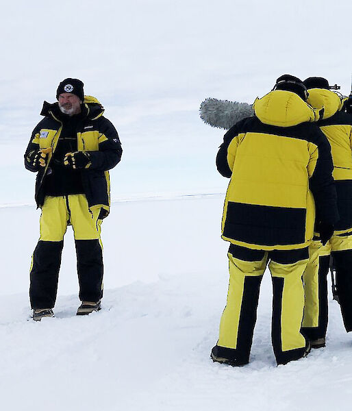 Yellow survival suited film crew on the ice with blue Hagglunds in near distance