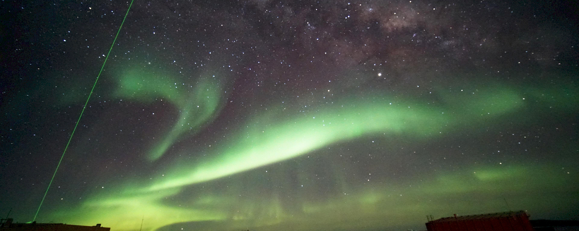 A green laser probes high-altitude clouds above Davis research station, in a night sky of auroras and stars.