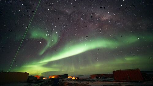 A green laser probes high-altitude clouds above Davis research station, in a night sky of auroras and stars.