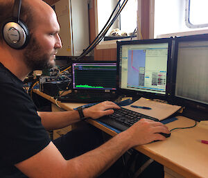 Dr Brian Miller listens for whales during a voyage, with a computer screen in front of him showing sounds.