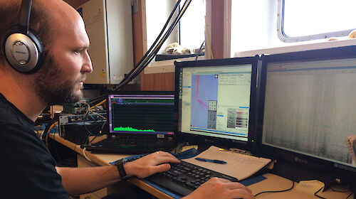 Dr Brian Miller listens for whales during a voyage, with a computer screen in front of him showing sounds.