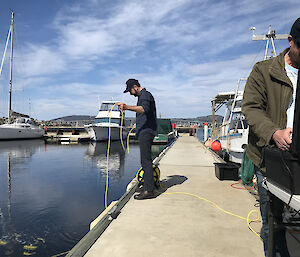 A man standing over water holding a yellow tether while another man stands at a black box with a computer in it.