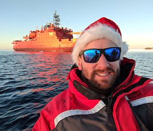 Brendan in a Santa hat in the foreground on a boat with Aurora Australis in the background