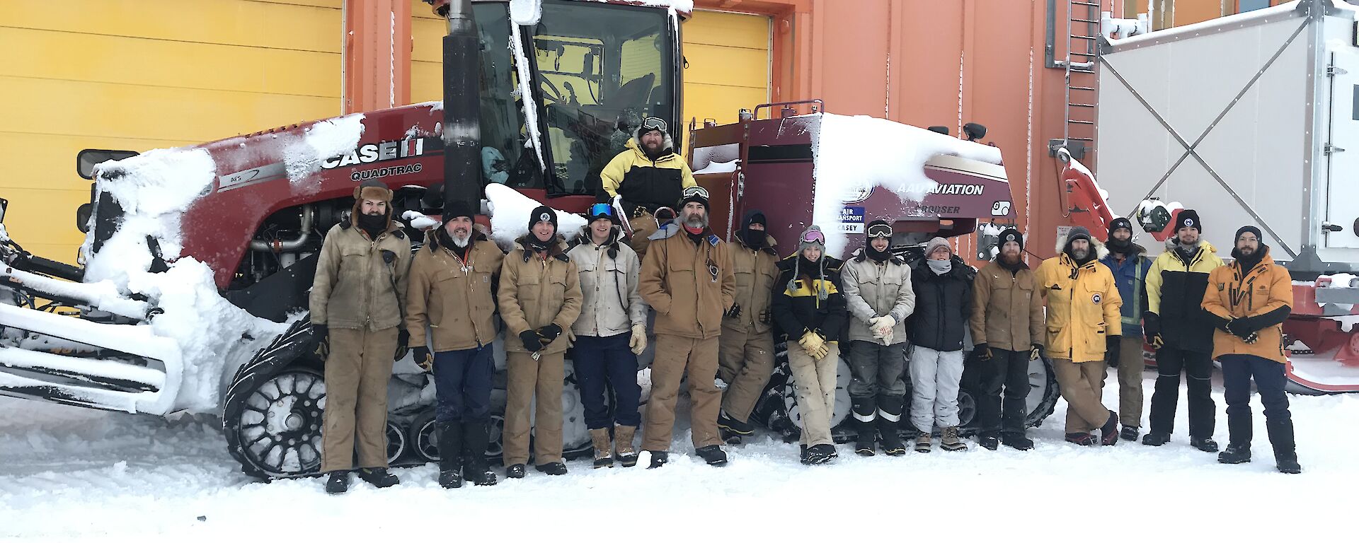 The 16 members of the traverse team standing in front of the red tracktor in front of the orange workshop