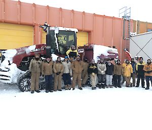 The 16 members of the traverse team standing in front of the red tracktor in front of the orange workshop