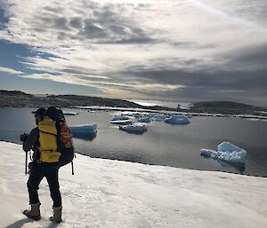 Juan walking with his field back with icebergs bits in the water in tha background