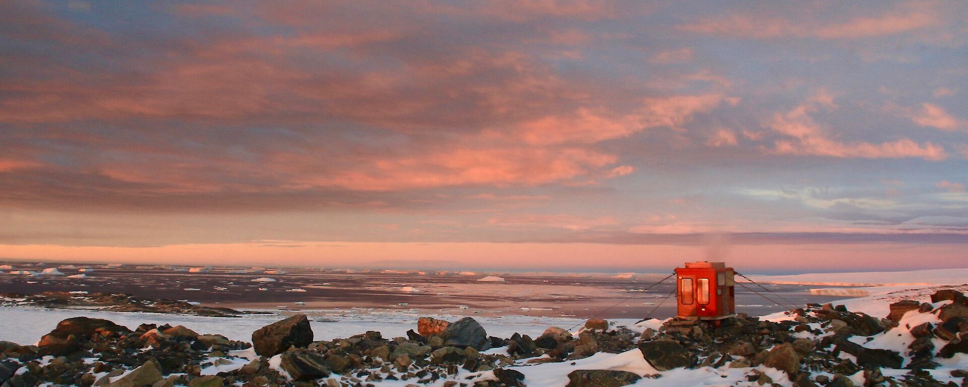 A red toilet building on rocks overlooking the bay at sunset
