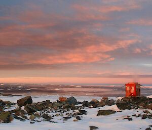 A red toilet building on rocks overlooking the bay at sunset