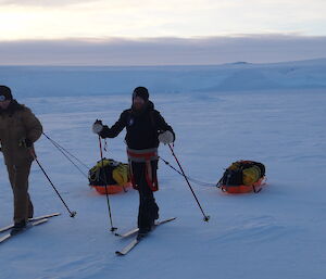 Two skiers towing sleds on the sea ice with ice cliffs in the background