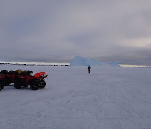 Thre quads stopped on the ice and one person standing in front a gigantic iceberg under a cloudy sky