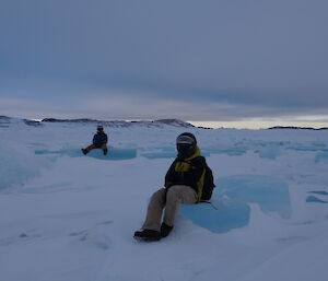 Two expeditioners in full winter dress and quad helmets sitting on large pieces of blue ice