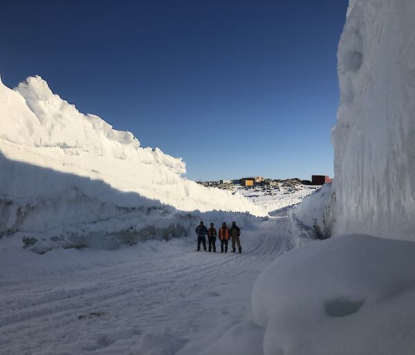 Four men on a snowy road with tall stacks of snow on either side of them.