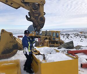 A yellow crane arm over a yellow skip bin filled with snow and a man wearing a blue jacket in front of the skip bin.