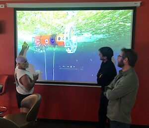 three men talking and pointing at a robot on a projector screen