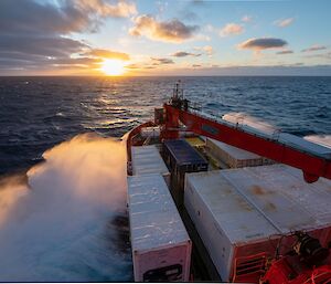 Sea spray covers the bow of a red ship, laden with white and blue containers, as it sails into a golden sunset across a windswept dark blue sea