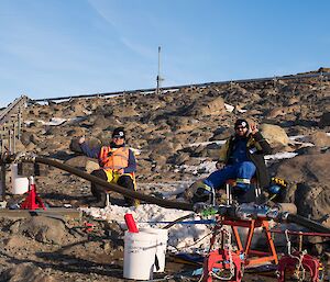 Two men in fold up chairs sit on a brown, rocky hill and keep watch over a fuel line valve assembly