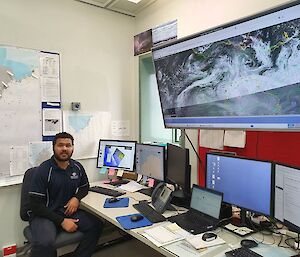 A man in blue casual office clothing sits before seven computer monitors as he creates weather forecasts using satellite images