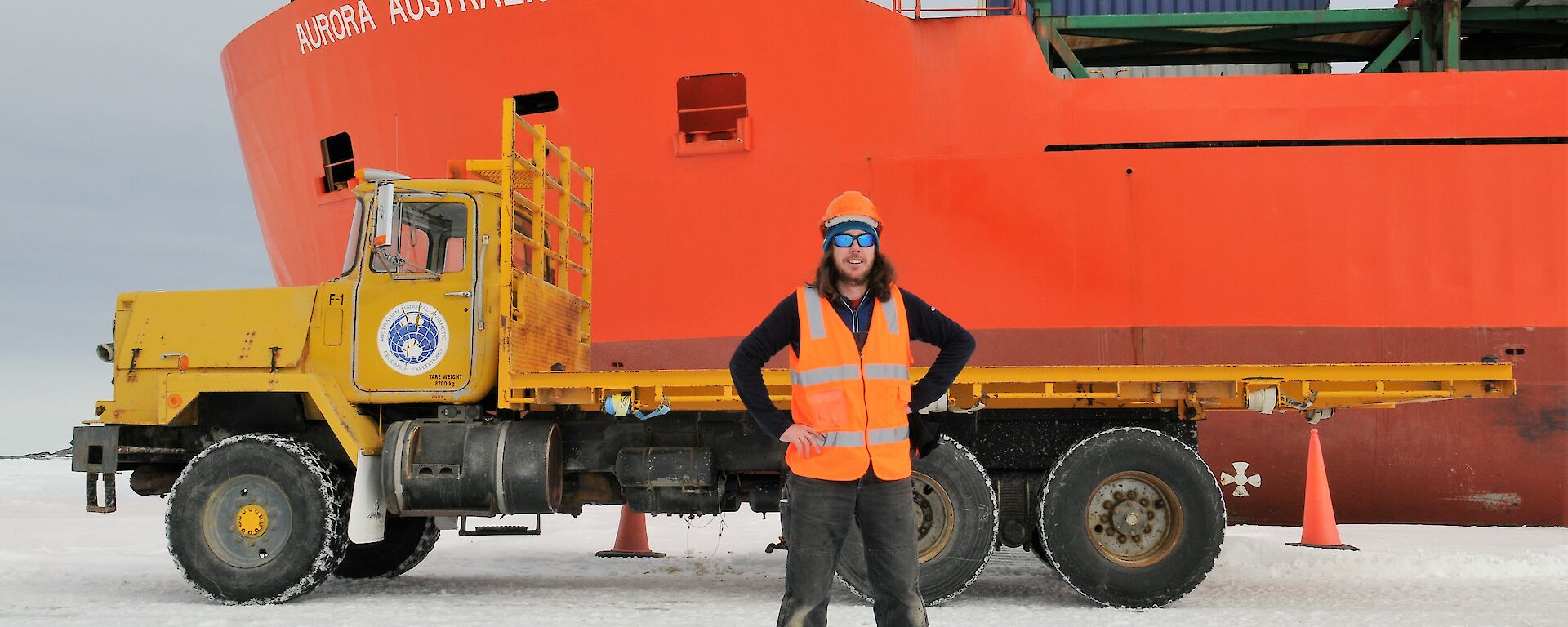 A man is standing on the sea ice in front of a yellow truck parked in front of a red icebreaker ship